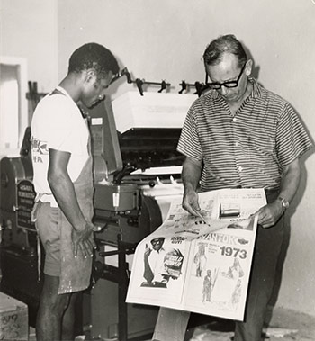 Two men looking at a newspaper next to a press